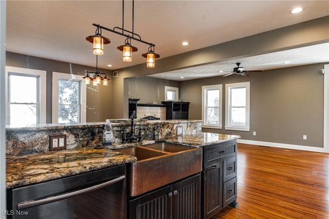 kitchen with stainless steel dishwasher, dark stone counters, ceiling fan with notable chandelier, wood-type flooring, and pendant lighting