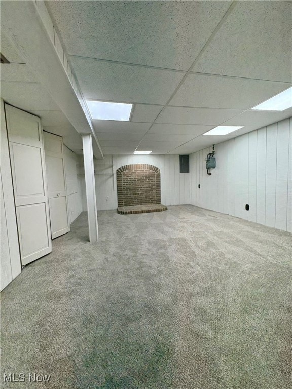 basement with a drop ceiling, carpet floors, wooden walls, and a brick fireplace