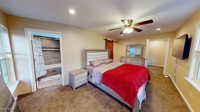 bedroom featuring a barn door, dark carpet, ceiling fan, and connected bathroom