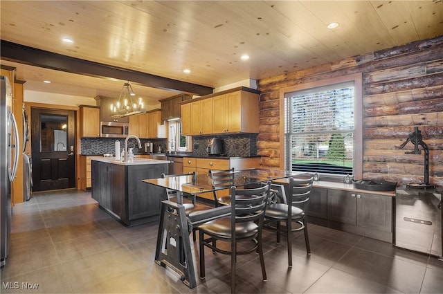 kitchen with appliances with stainless steel finishes, tasteful backsplash, log walls, hanging light fixtures, and an island with sink