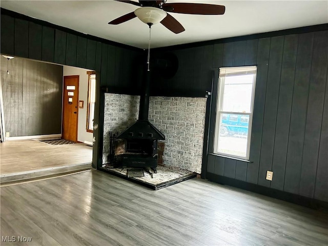 unfurnished living room featuring a wood stove, wooden walls, light hardwood / wood-style flooring, and ceiling fan