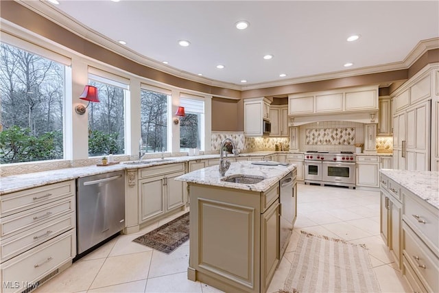 kitchen featuring appliances with stainless steel finishes, light stone counters, a kitchen island with sink, sink, and cream cabinets