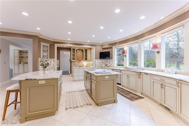 kitchen with cream cabinets, ornamental molding, light stone countertops, an island with sink, and tasteful backsplash