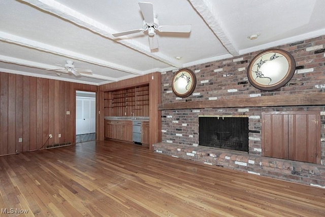 unfurnished living room featuring wood walls, a brick fireplace, ceiling fan, beamed ceiling, and wood-type flooring