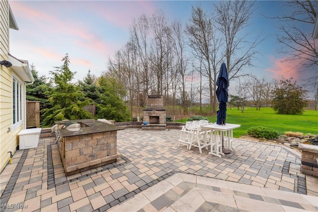 patio terrace at dusk featuring an outdoor stone fireplace and a lawn