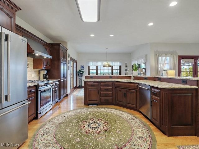 kitchen with high quality appliances, wall chimney exhaust hood, decorative backsplash, decorative light fixtures, and light hardwood / wood-style floors