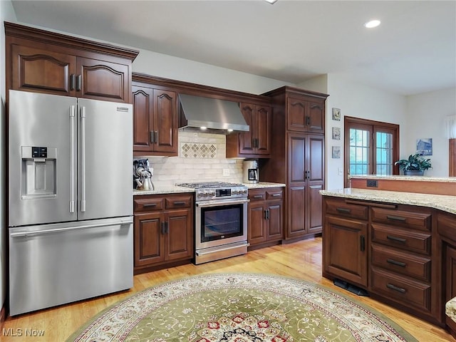 kitchen featuring light stone countertops, tasteful backsplash, stainless steel appliances, wall chimney range hood, and light hardwood / wood-style flooring