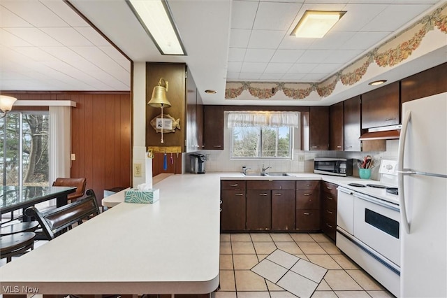 kitchen with a breakfast bar, white appliances, sink, wooden walls, and light tile patterned flooring