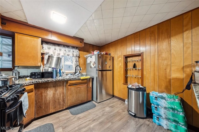 kitchen featuring stainless steel refrigerator, stone counters, black range with gas stovetop, wood walls, and light hardwood / wood-style floors