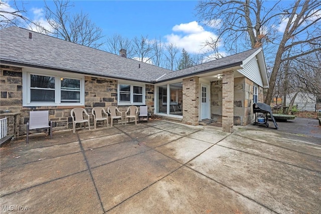 rear view of house featuring a chimney, a patio, roof with shingles, and stone siding