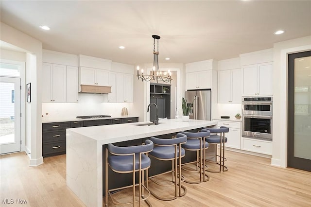 kitchen featuring pendant lighting, a kitchen island with sink, white cabinets, light wood-type flooring, and appliances with stainless steel finishes