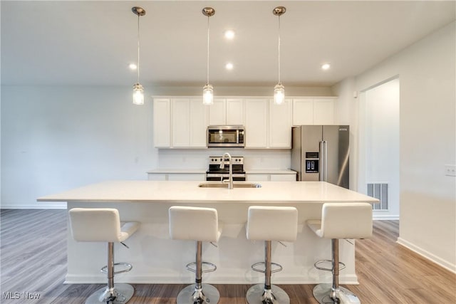 kitchen with appliances with stainless steel finishes, decorative light fixtures, light hardwood / wood-style flooring, a center island with sink, and white cabinetry