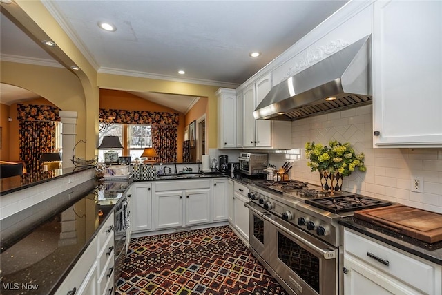 kitchen with wall chimney exhaust hood, dark stone countertops, range with two ovens, decorative backsplash, and white cabinets