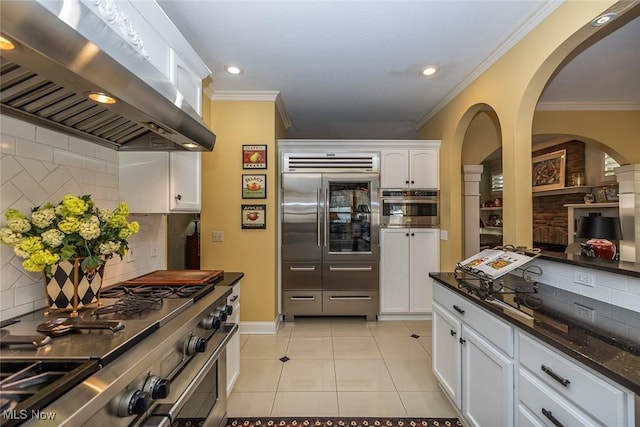 kitchen featuring wall chimney range hood, high quality appliances, dark stone counters, decorative backsplash, and white cabinets