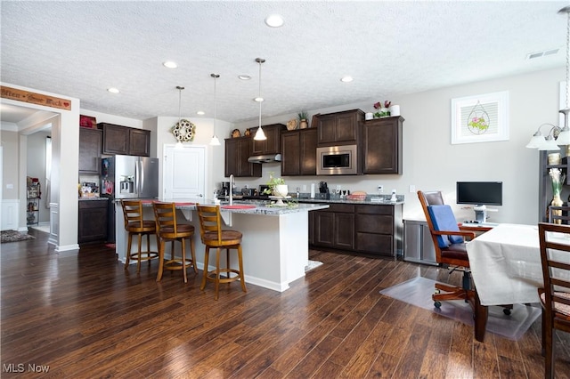 kitchen with a kitchen bar, appliances with stainless steel finishes, decorative light fixtures, dark hardwood / wood-style floors, and an island with sink