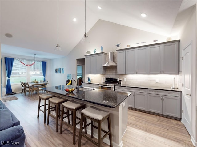 kitchen featuring a kitchen island with sink, high vaulted ceiling, wall chimney range hood, hanging light fixtures, and stainless steel range oven