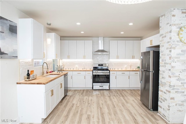 kitchen featuring butcher block counters, sink, wall chimney range hood, white cabinets, and appliances with stainless steel finishes