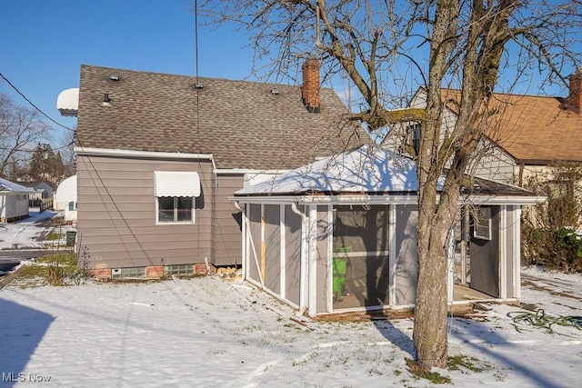 snow covered rear of property featuring a sunroom, a chimney, and a shingled roof