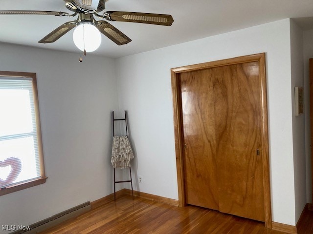 unfurnished bedroom featuring hardwood / wood-style flooring, ceiling fan, multiple windows, and a baseboard heating unit