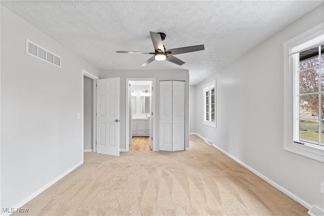 unfurnished bedroom with light carpet, ensuite bathroom, ceiling fan, a textured ceiling, and a closet