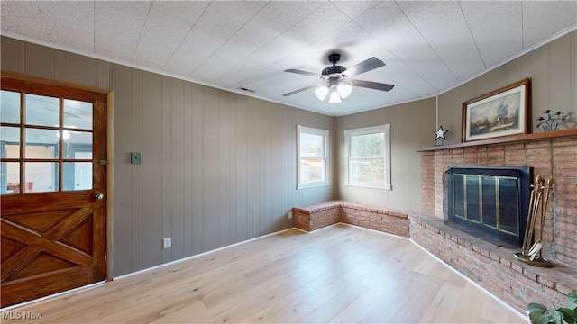 unfurnished living room with crown molding, wooden walls, a brick fireplace, ceiling fan, and light hardwood / wood-style floors