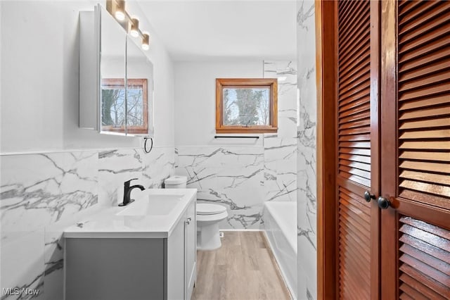 bathroom featuring a tub to relax in, wood-type flooring, toilet, vanity, and tile walls