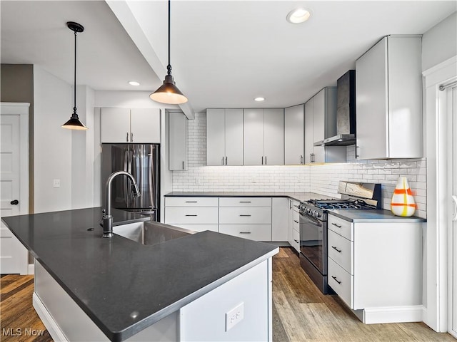 kitchen featuring a kitchen island with sink, wall chimney exhaust hood, gas range oven, decorative light fixtures, and stainless steel refrigerator