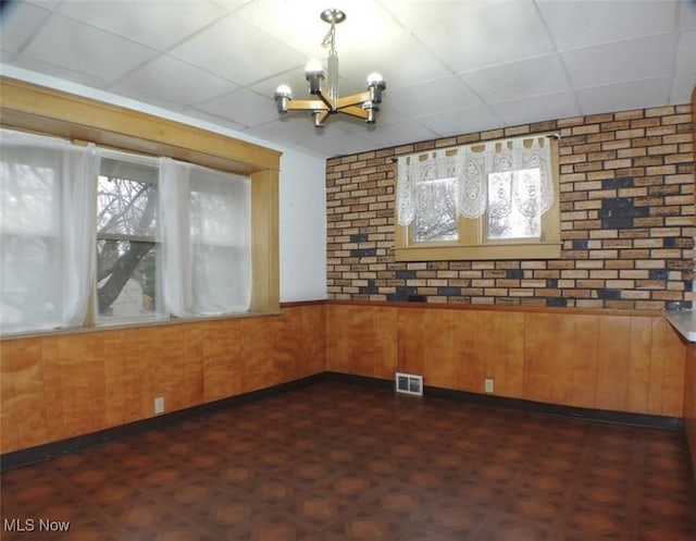 empty room featuring dark parquet flooring, a drop ceiling, brick wall, wooden walls, and a chandelier