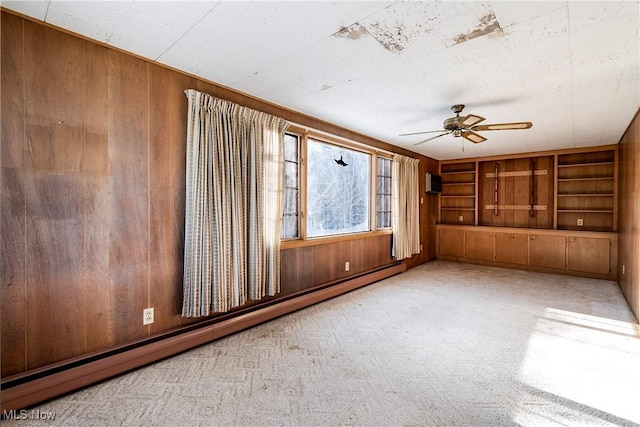 spare room featuring built in shelves, ceiling fan, a baseboard radiator, wood walls, and light colored carpet