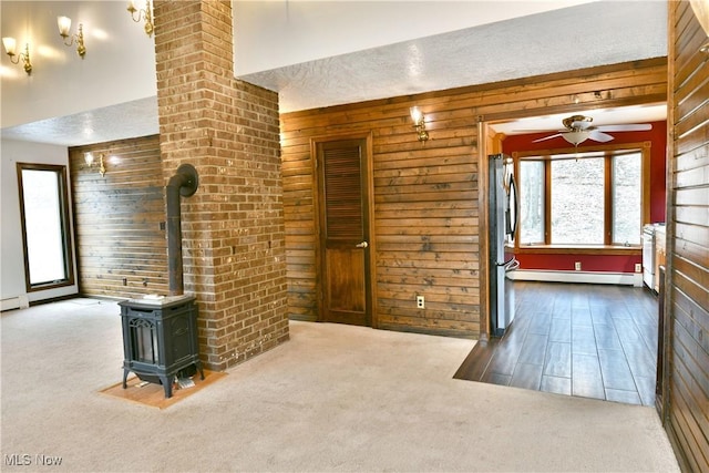 unfurnished living room featuring a wood stove, ceiling fan, a baseboard heating unit, carpet floors, and a textured ceiling