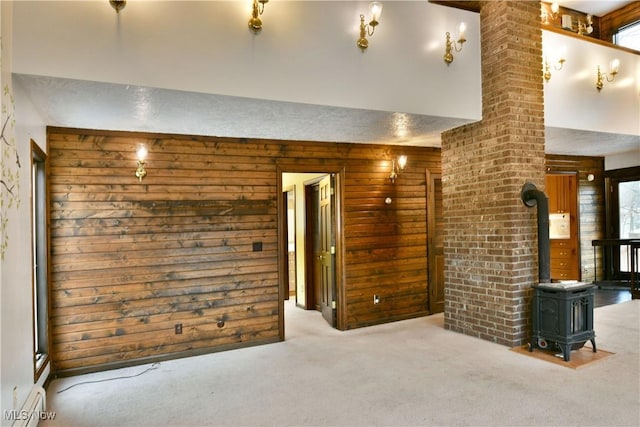 empty room featuring light colored carpet, a healthy amount of sunlight, wooden walls, a baseboard radiator, and a wood stove