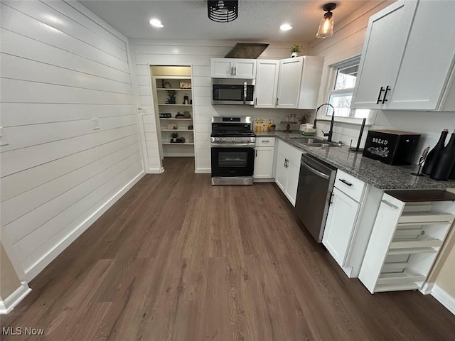 kitchen with appliances with stainless steel finishes, wooden walls, sink, dark stone countertops, and white cabinetry