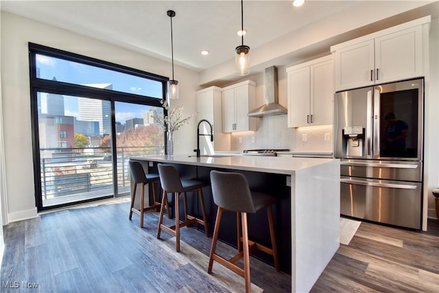 kitchen featuring white cabinets, a kitchen island with sink, stainless steel fridge with ice dispenser, and wall chimney range hood