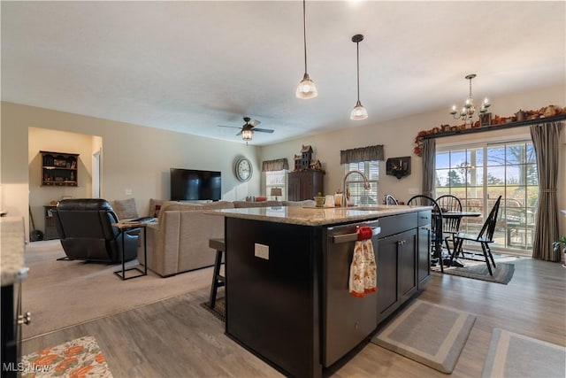 kitchen with light stone countertops, hanging light fixtures, stainless steel dishwasher, a center island with sink, and ceiling fan with notable chandelier