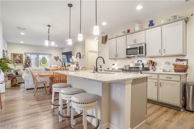 kitchen with sink, stainless steel appliances, light stone counters, an island with sink, and pendant lighting