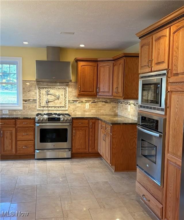 kitchen with wall chimney exhaust hood, stainless steel appliances, dark stone counters, and tasteful backsplash