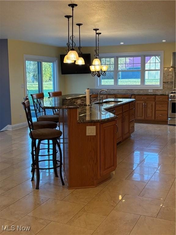 kitchen featuring sink, electric range, decorative light fixtures, dark stone countertops, and an island with sink