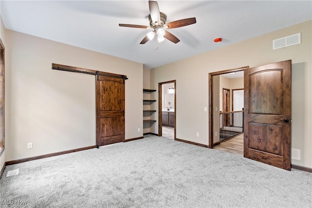 unfurnished bedroom featuring a barn door, ensuite bathroom, ceiling fan, and light colored carpet