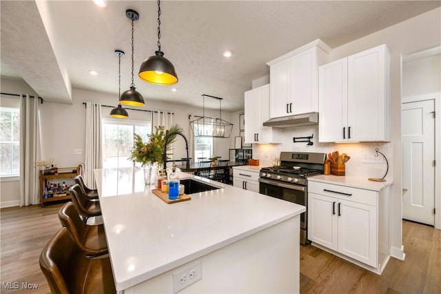 kitchen featuring gas stove, a kitchen island with sink, sink, white cabinetry, and hanging light fixtures