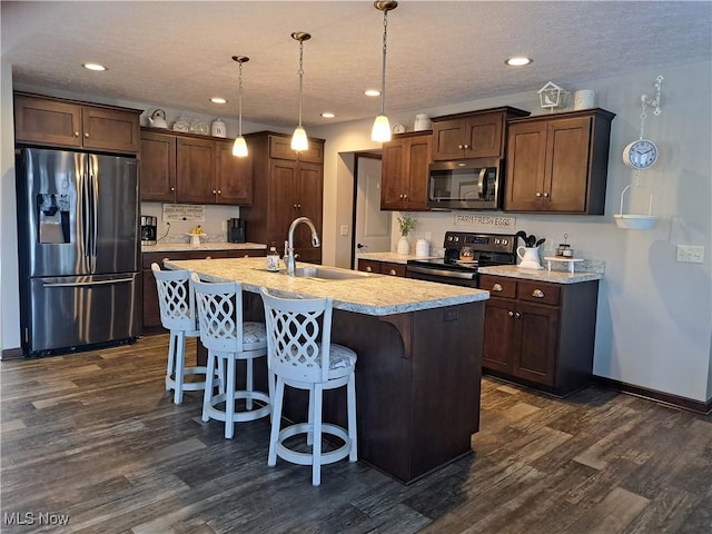 kitchen featuring sink, dark wood-type flooring, decorative light fixtures, a center island with sink, and appliances with stainless steel finishes