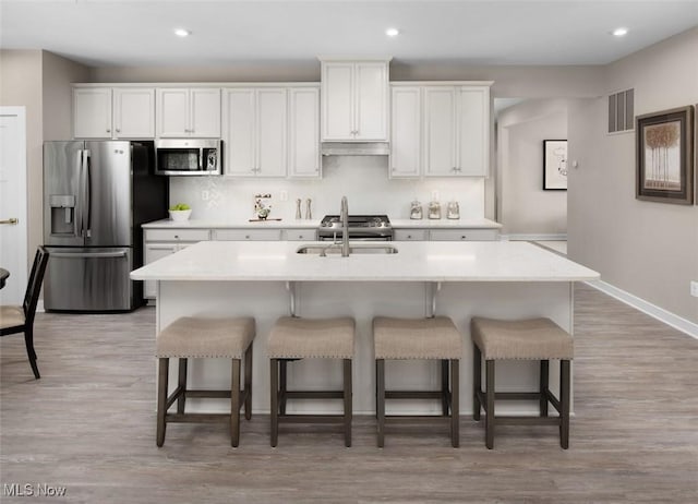 kitchen with white cabinets, appliances with stainless steel finishes, light hardwood / wood-style floors, and a kitchen island with sink