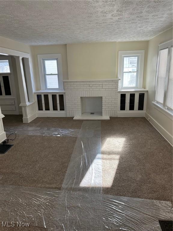 unfurnished living room with a wealth of natural light, dark colored carpet, a textured ceiling, and a brick fireplace