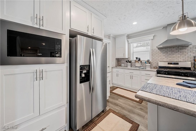 kitchen with pendant lighting, white cabinets, wall chimney exhaust hood, light wood-type flooring, and appliances with stainless steel finishes