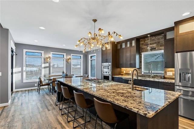 kitchen with appliances with stainless steel finishes, hanging light fixtures, dark brown cabinetry, light stone countertops, and a center island with sink