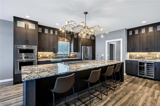 kitchen featuring appliances with stainless steel finishes, an island with sink, beverage cooler, light stone counters, and dark brown cabinetry