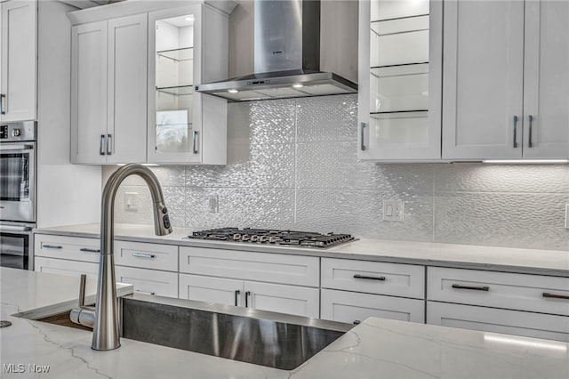 kitchen with tasteful backsplash, light stone counters, sink, wall chimney range hood, and white cabinets