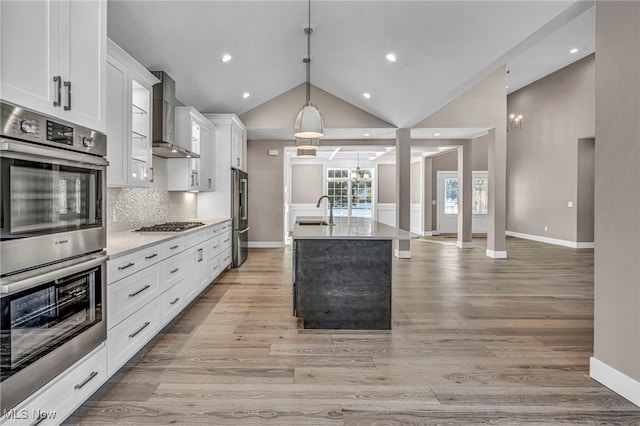 kitchen with sink, stainless steel appliances, wall chimney range hood, a kitchen island with sink, and white cabinets