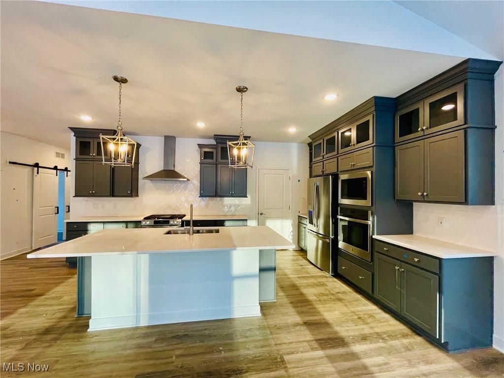 kitchen with a barn door, decorative backsplash, appliances with stainless steel finishes, wall chimney range hood, and a sink