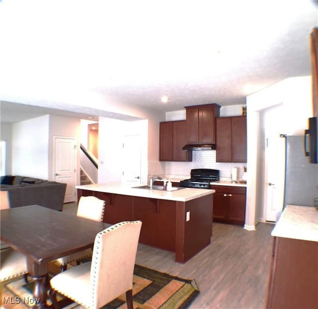 kitchen featuring dark wood-type flooring, a center island with sink, black gas stove, sink, and decorative backsplash