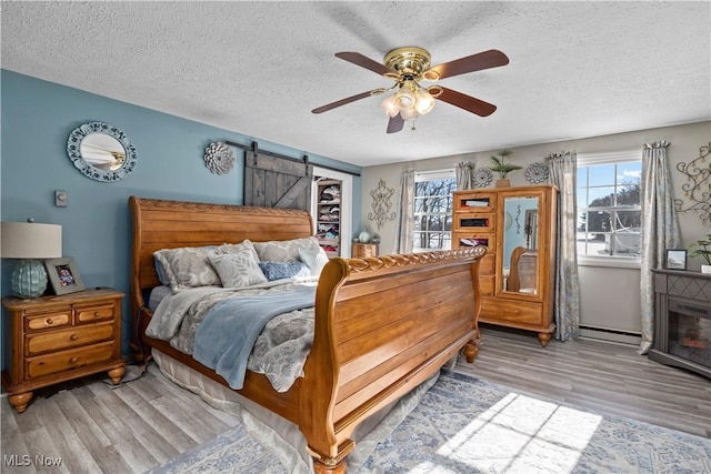 bedroom featuring a textured ceiling, ceiling fan, a barn door, a baseboard radiator, and light hardwood / wood-style flooring